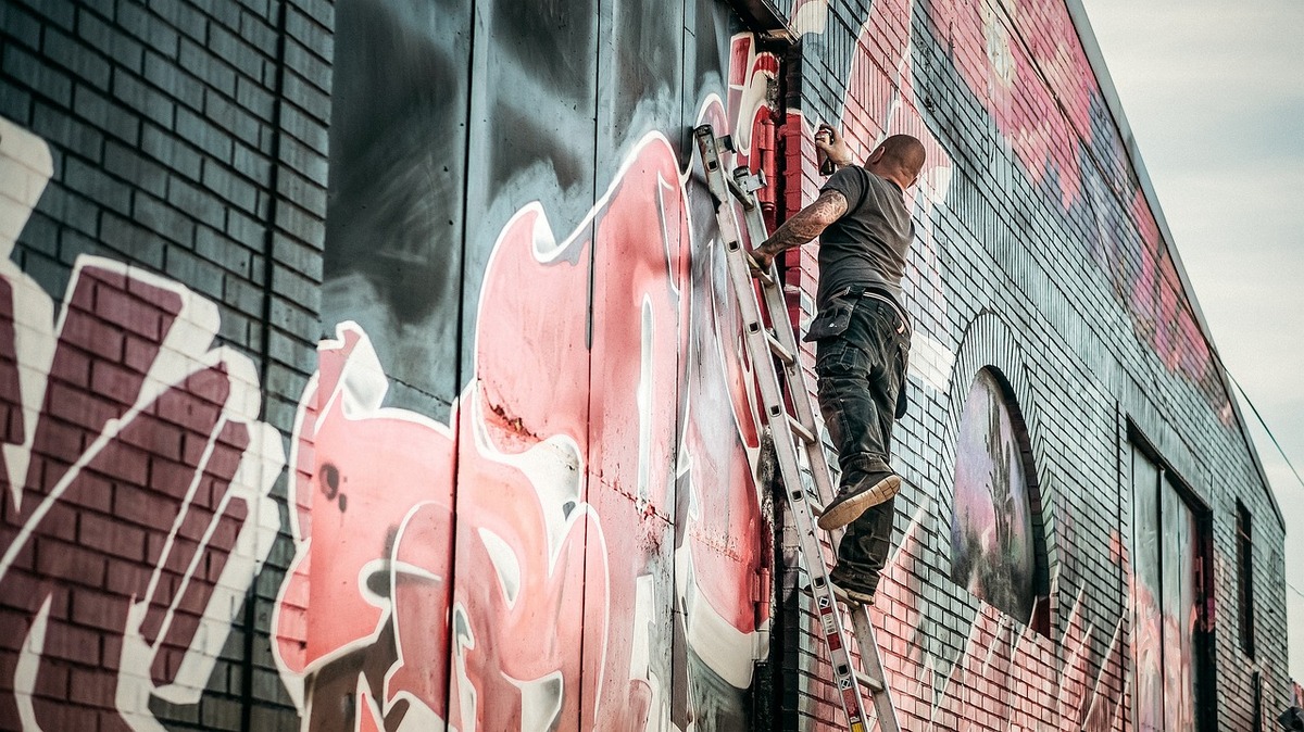 Ein Graffitti Künstler sprayed eine Wand an. er steht auf einer Leiter.